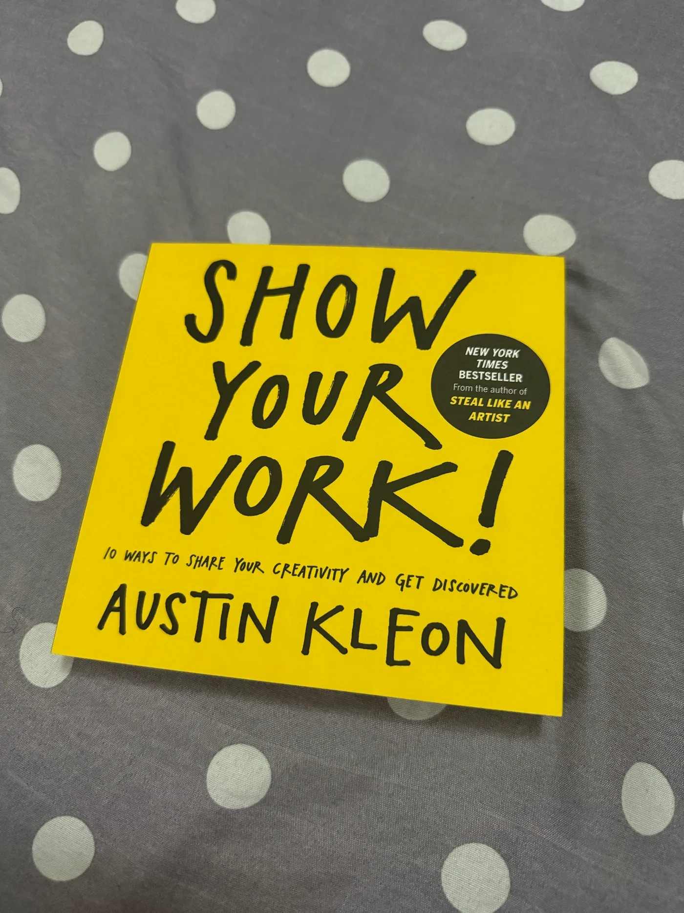 A yellow paperback copy of Show Your Work! by Austin Kleon lying on a grey bedsheet with white polka dots.