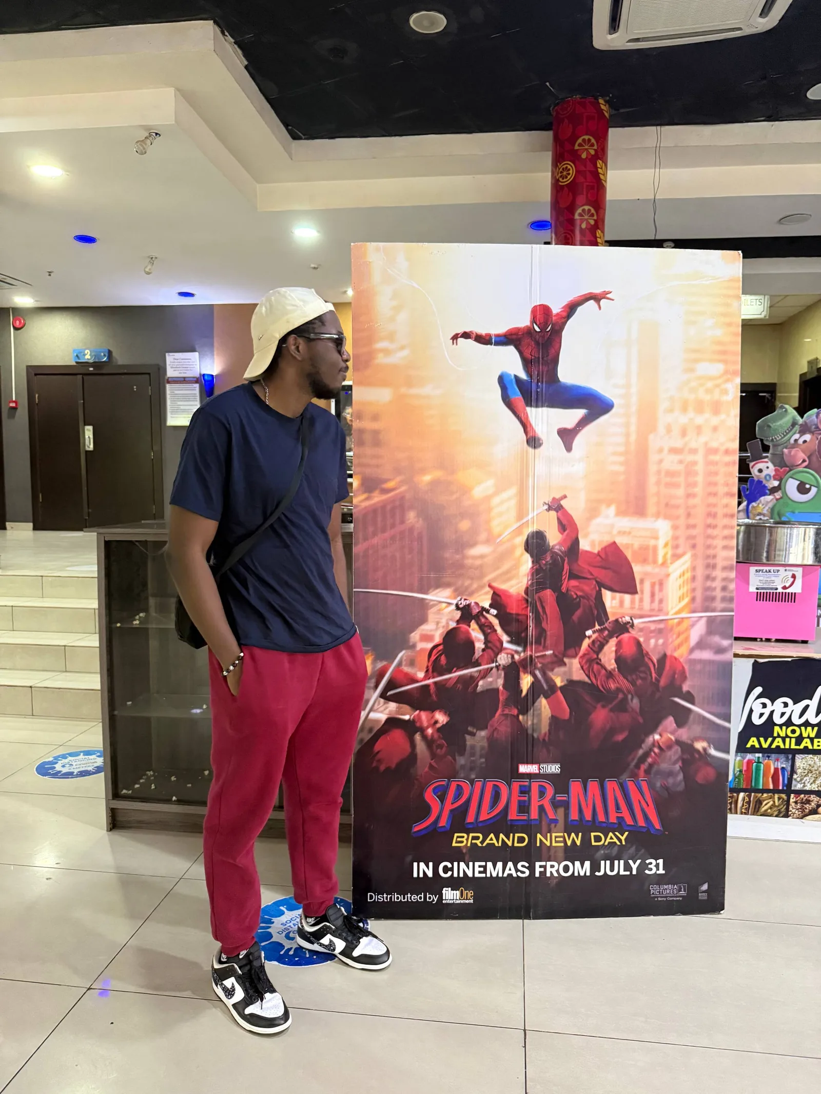 A man in a cap and red trousers beside a large Spider-Man: Brand New Day standee in a cinema lobby.