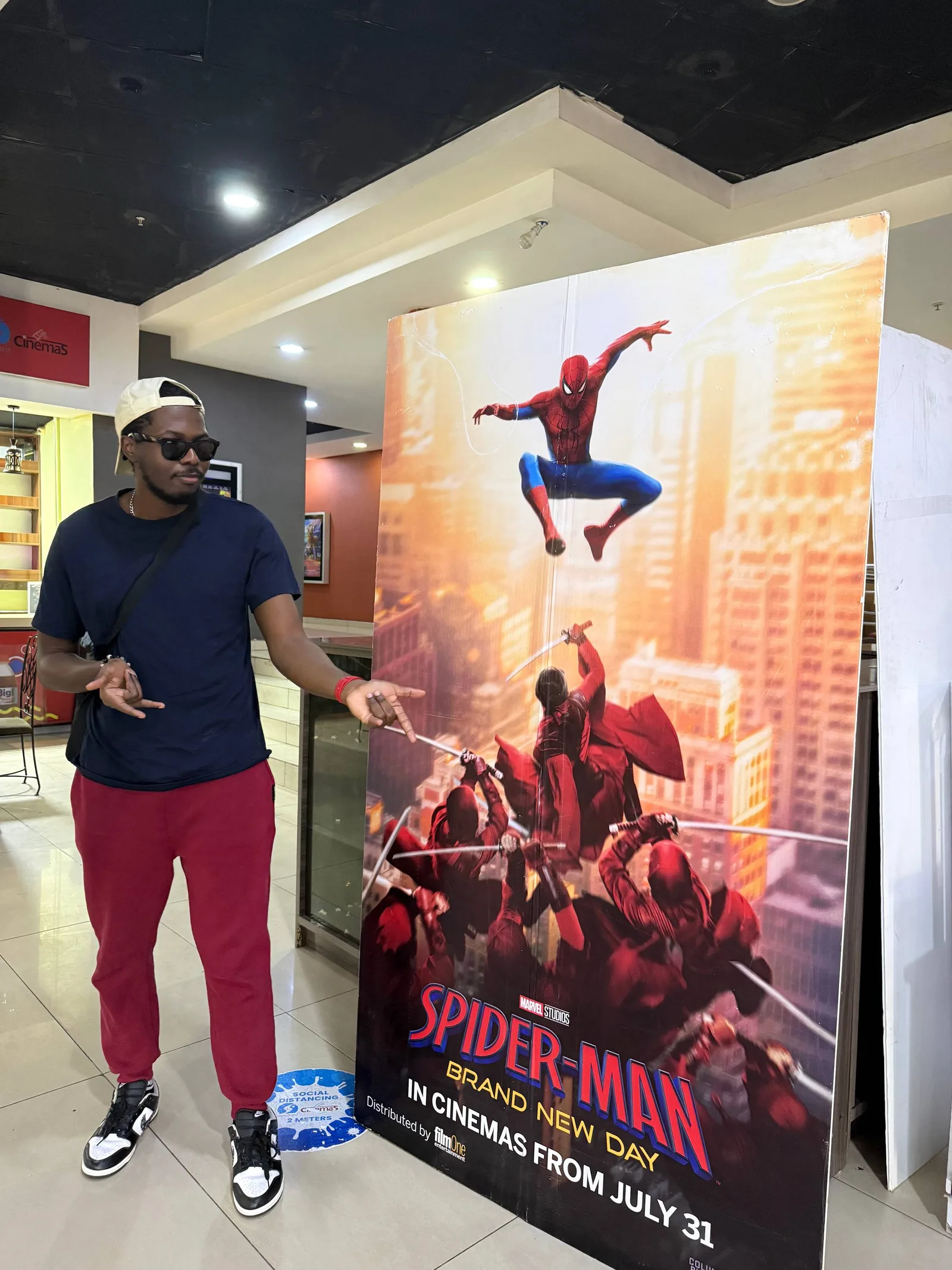 A man in sunglasses and red trousers beside a Spider-Man: Brand New Day standee, one hand reaching towards it.